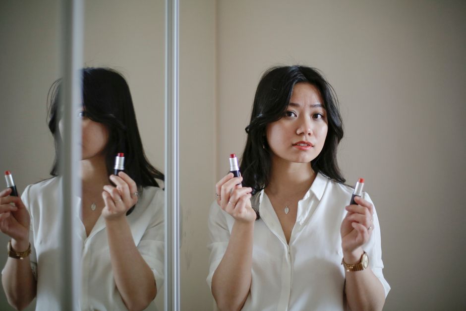 Young Asian woman looking at camera and selecting between lipsticks while standing near mirror during makeup application at home