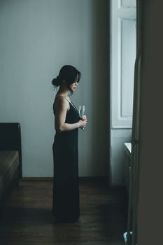 一位悲伤的亚洲女性身穿优雅的黑色连衣裙，侧身站在家中昏暗的房间里，手里拿着一杯酒