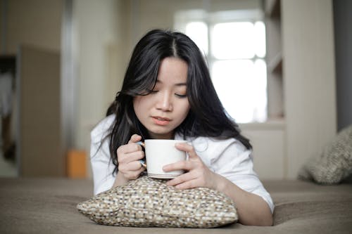 Young ethnic female enjoying hot drink and lying on soft bed in calm morning at home