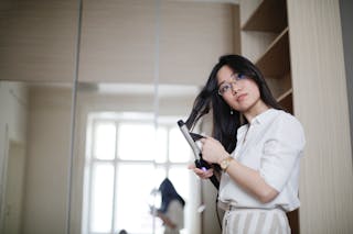 Low angle of stylish Asian female curling hair with iron and looking away in morning at home