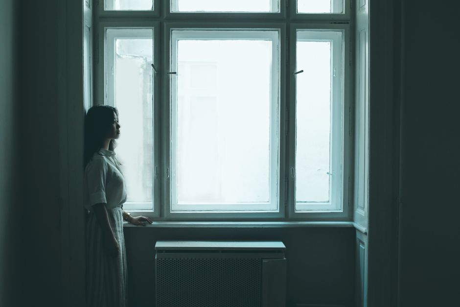 어두운 방 안에서 젊은 여성이 크게 창가에 서서 사색에 잠겨 있는 모습으로, 고독을 반영합니다.