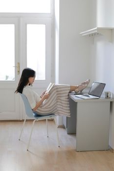 Side view of young Asian female student keeping legs on table near laptop and reading textbook in cozy room at home