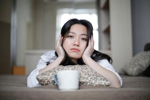 Young Asian female lying on soft pillow on comfortable bed and looking at camera in morning at home