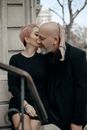 Elegant adult female with stylish short haircut embracing and kissing gently bald bearded man near stone building in city