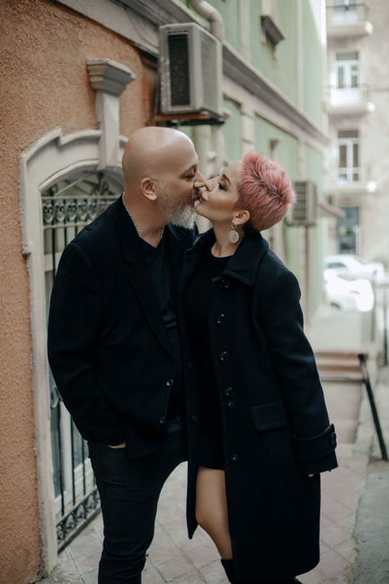 Side view of stylish bearded bald mature man and elegant woman in black outfits kissing tenderly while standing near old stone building on city street