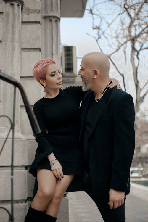 Free Elegant short haired woman in black dress embracing stylish bald middle aged man while standing together near old stone building in city Stock Photo