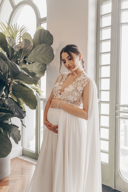 Peaceful tender young pregnant female in stylish vintage white lace dress touching belly while standing in light room of old building