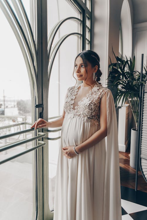 Young pregnant woman in vintage gown looking at window