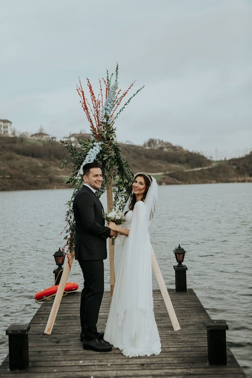 Full body side view of happy young newlywed couple in elegant wedding clothes holding hands and looking at camera while standing on wooden pier decorated with plants at riverside
