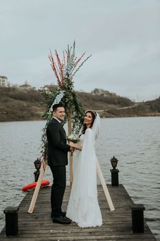 一对幸福的年轻新婚夫妇身着优雅的婚礼服装，手牵手站在河边装饰着植物的木墩上，面向镜头拍摄全身侧视图