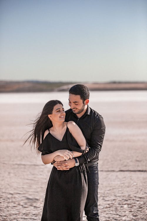 Cheerful young couple in stylish black clothes hugging and laughing while enjoying romantic moments together at seaside