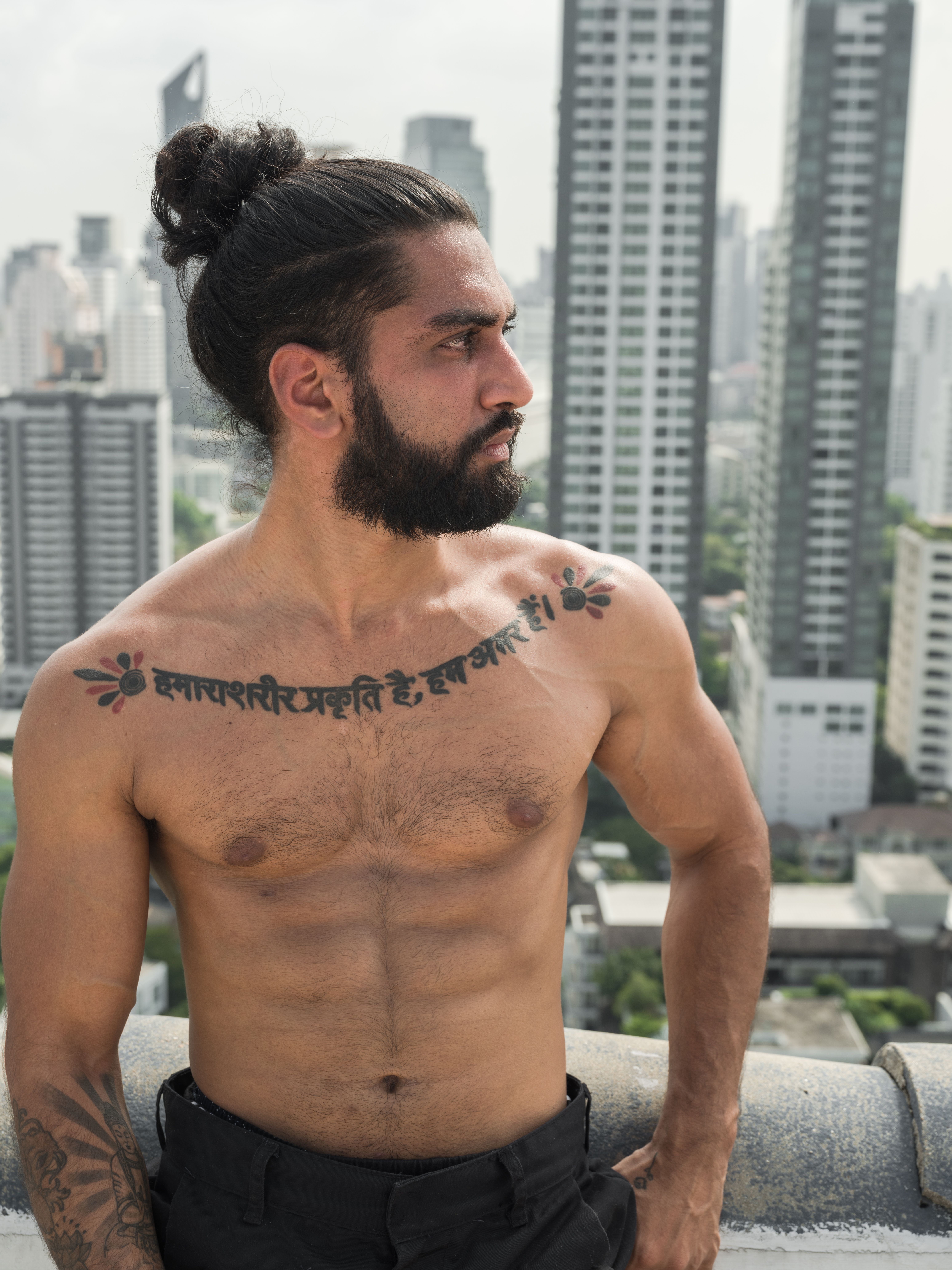 Shirtless muscular man with tattoos gazing away against an urban skyline view.