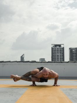 A fit man doing yoga pose on rooftop helipad, showcasing strength and flexibility against urban skyline.