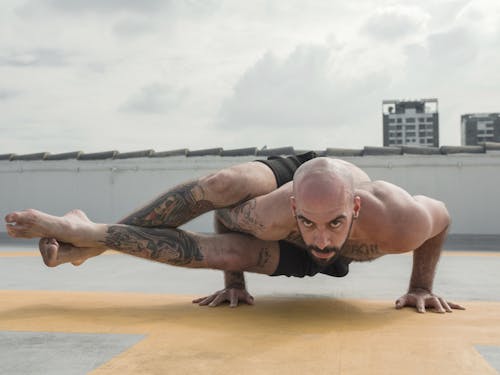 Full length shirtless brutal bearded tattooed male doing eight angle pose while practicing yoga on roof against cloudy sky