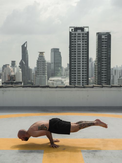 Free Shirtless man practicing advanced yoga pose on rooftop helipad with city skyline background. Stock Photo