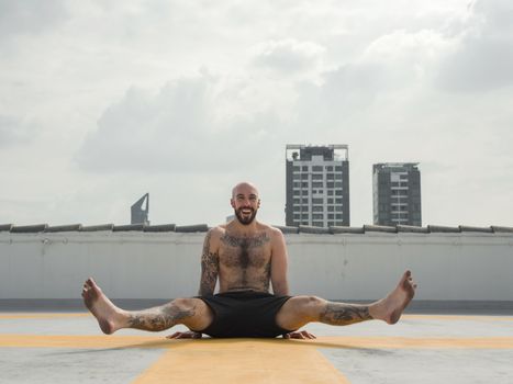 Tattooed man performing yoga pose on rooftop with city skyline in background.