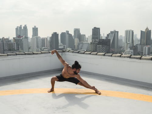 Full length of muscular shirtless male practicing martial arts on rooftop against modern skyscrapers on cloudy day