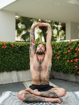 A fit tattooed man practicing yoga with arms raised in an outdoor setting, exemplifying mindfulness and balance.