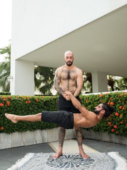 Two shirtless men performing acro yoga with focus and strength, set in an outdoor garden.
