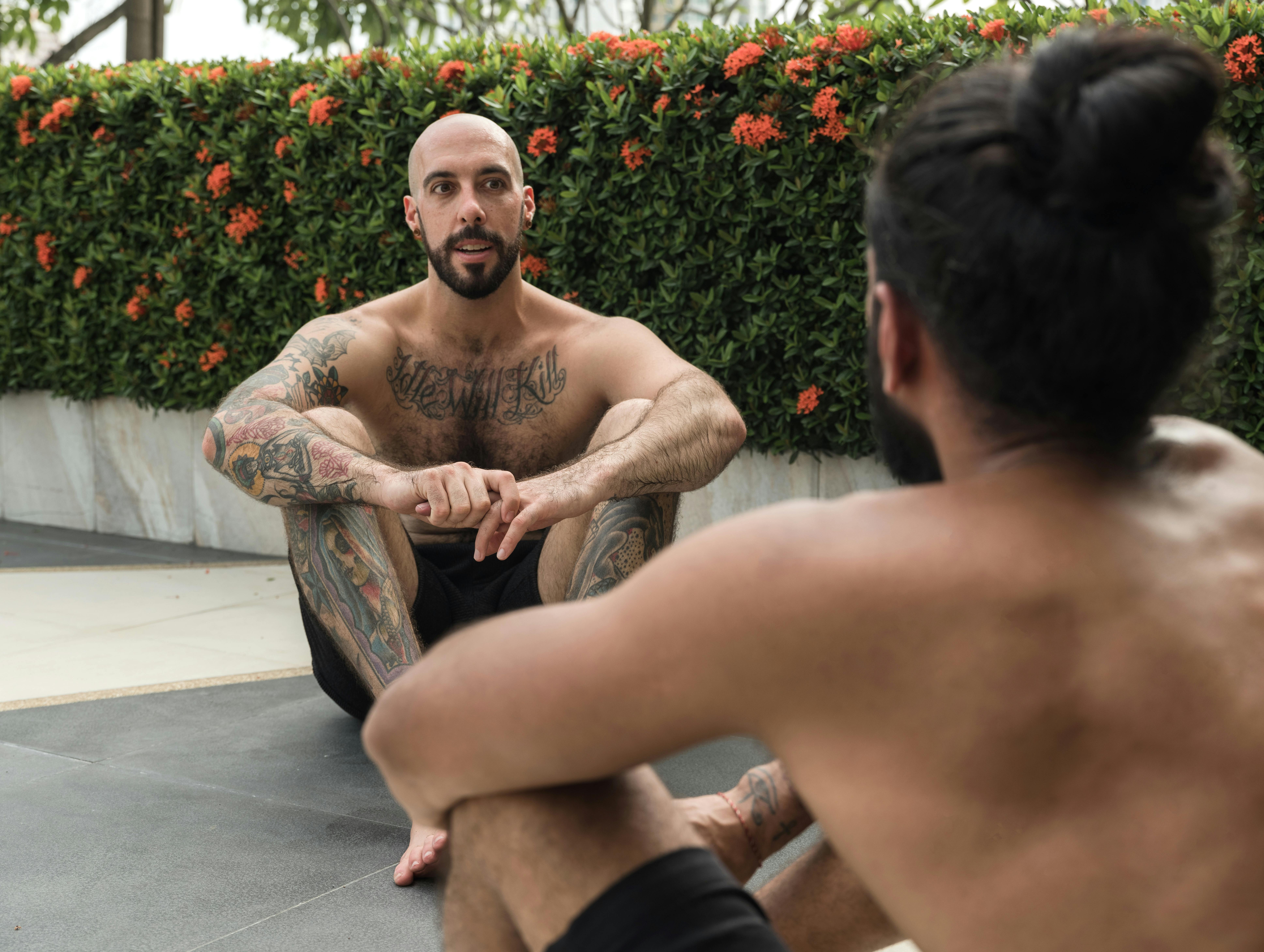 Two men having a casual, shirtless conversation outdoors amidst greenery.