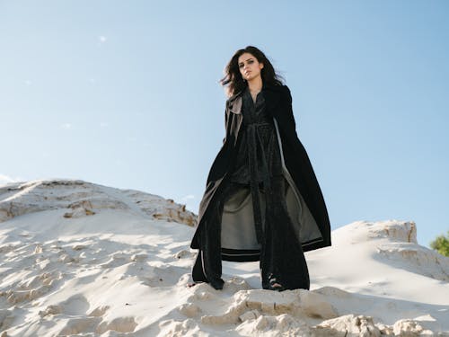 From below full length young pensive good looking brunette in trendy black outerwear with hands in pocket standing on sandy cliff against blue cloudless sky and looking at camera