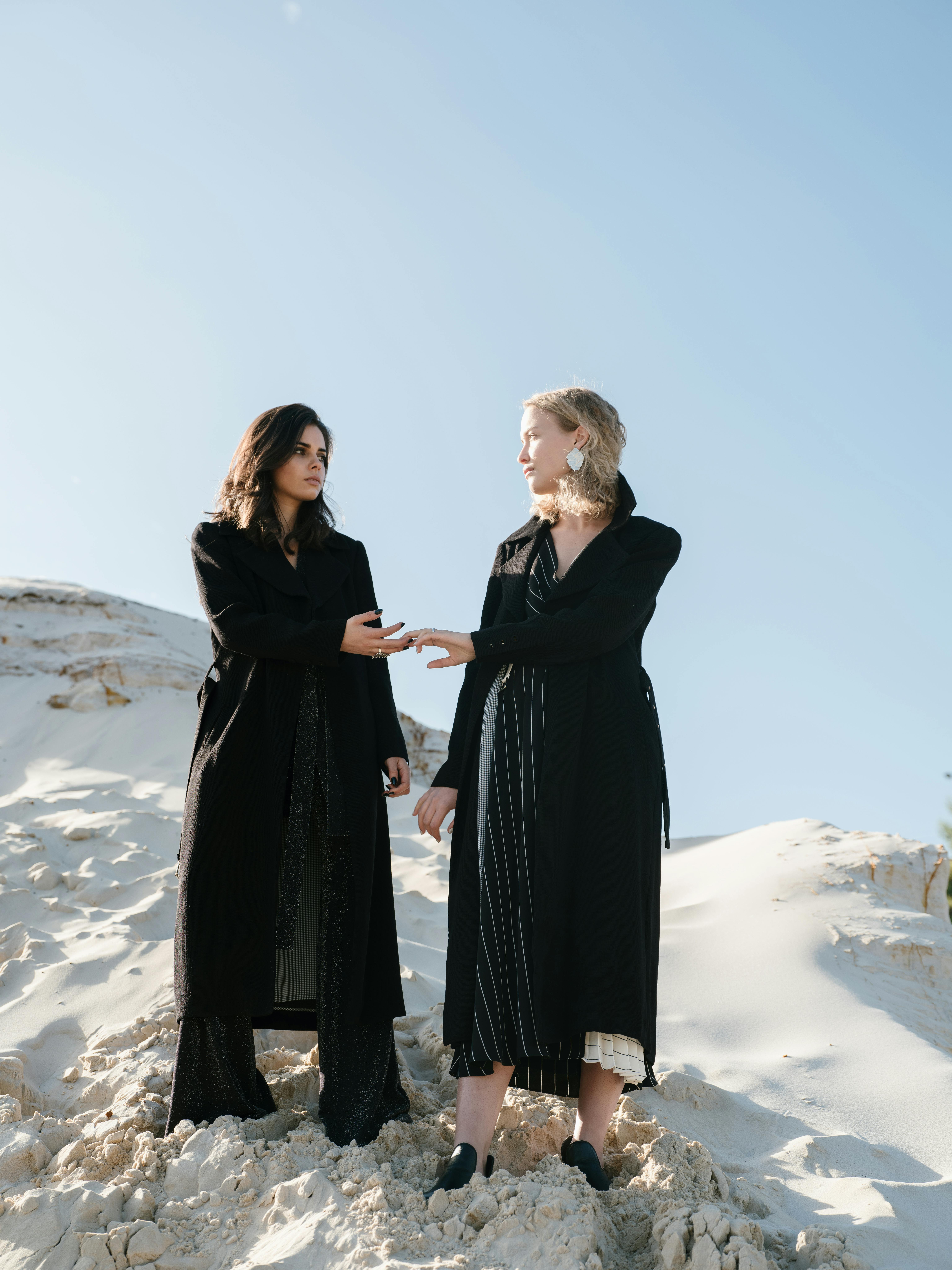 From below of young calm females in stylish black clothes holding hands and looking at each other while standing on white sandy slope against blue cloudless sky