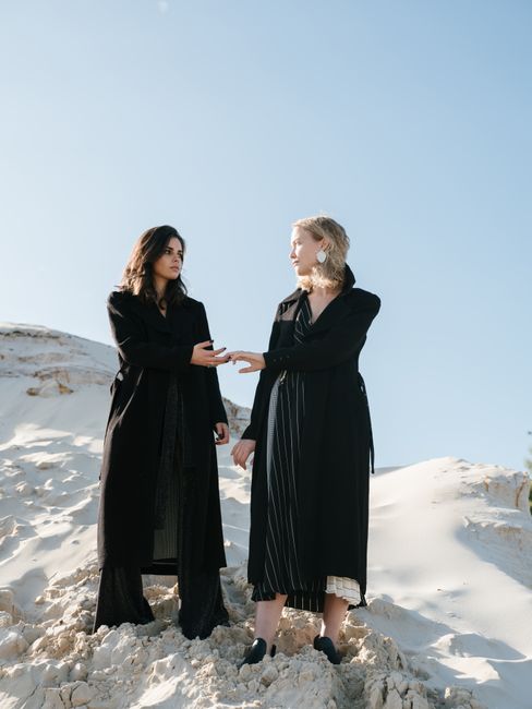From below of young calm females in stylish black clothes holding hands and looking at each other while standing on white sandy slope against blue cloudless sky