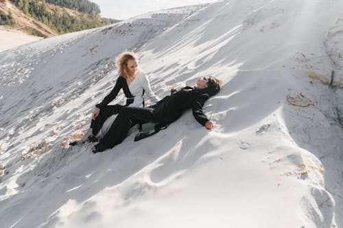 Full body unrecognizable calm females in stylish black and white outfits resting on white sand near green hills on sunny weather