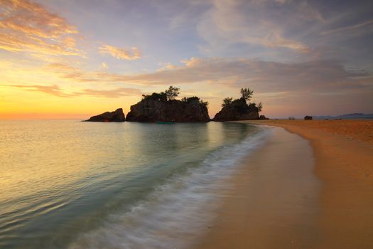 Tranquil beach scene at sunset with gentle waves and a scenic rocky outcrop.