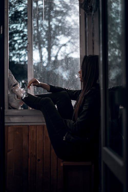 Side view of unrecognizable calm lonely female teenager sitting near open window in dark room and looking away pensively
