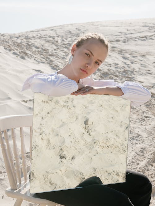 Pensive woman with square mirror sitting near sandy hill