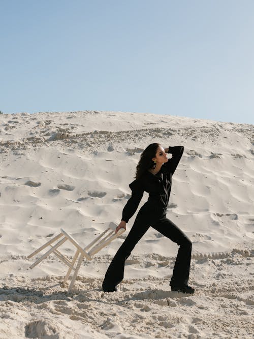 Full length of attractive brunette with long hair wearing black outfit posing with white wooden stool on white sand under clear blue sky during daytime while looking away