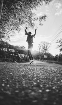 Black and white low angle of anonymous active teenager jumping with arms raised on paved alley in park in sunny day