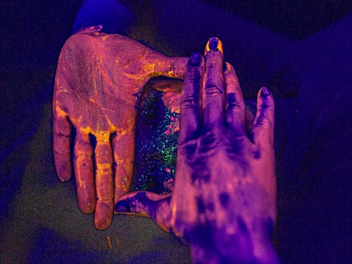 Unrecognizable female covering painted face with hands covered with colorful pigment in dark studio with neon illumination