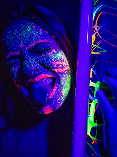 Crop grimacing female with glowing multicolored paints on face holding black light lamp and keeping tongue out