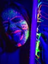 Crop grimacing female with glowing multicolored paints on face holding black light lamp and keeping tongue out