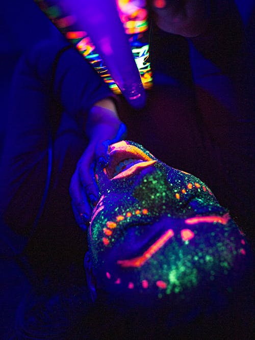 Free From above seductive female with glowing multicolored neon paints on face lying on floor with mouth opened sensually while touching chin and holding UV lamp Stock Photo
