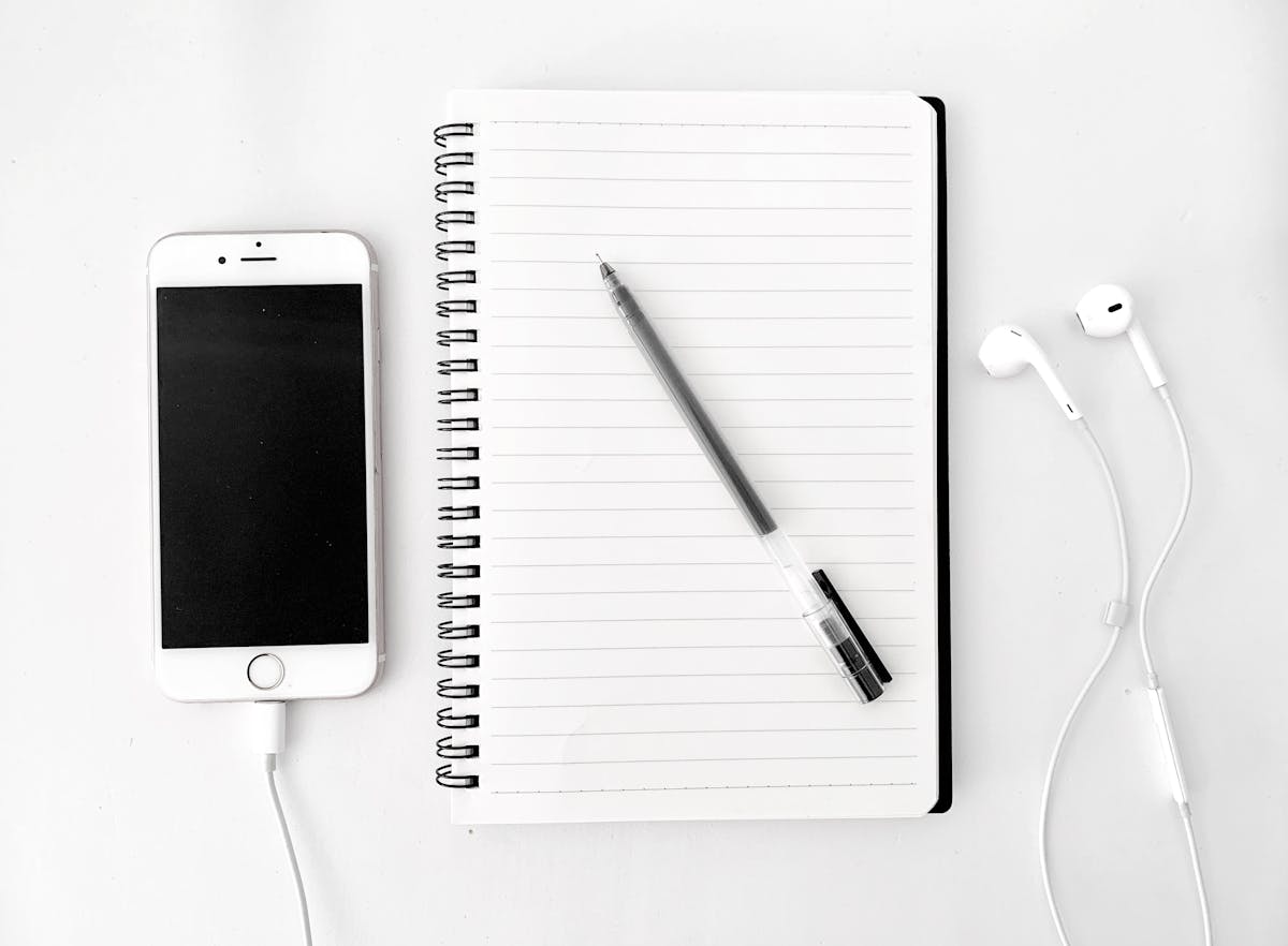 Top view arrangement of spiral notebook with blank sheets and pen placed on white desk near modern mobile phone with earphones