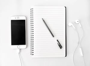 Top view arrangement of spiral notebook with blank sheets and pen placed on white desk near modern mobile phone with earphones