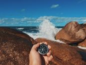 Crop anonymous male tourist with compass in hand standing on massive stony shore washed by splashing waves
