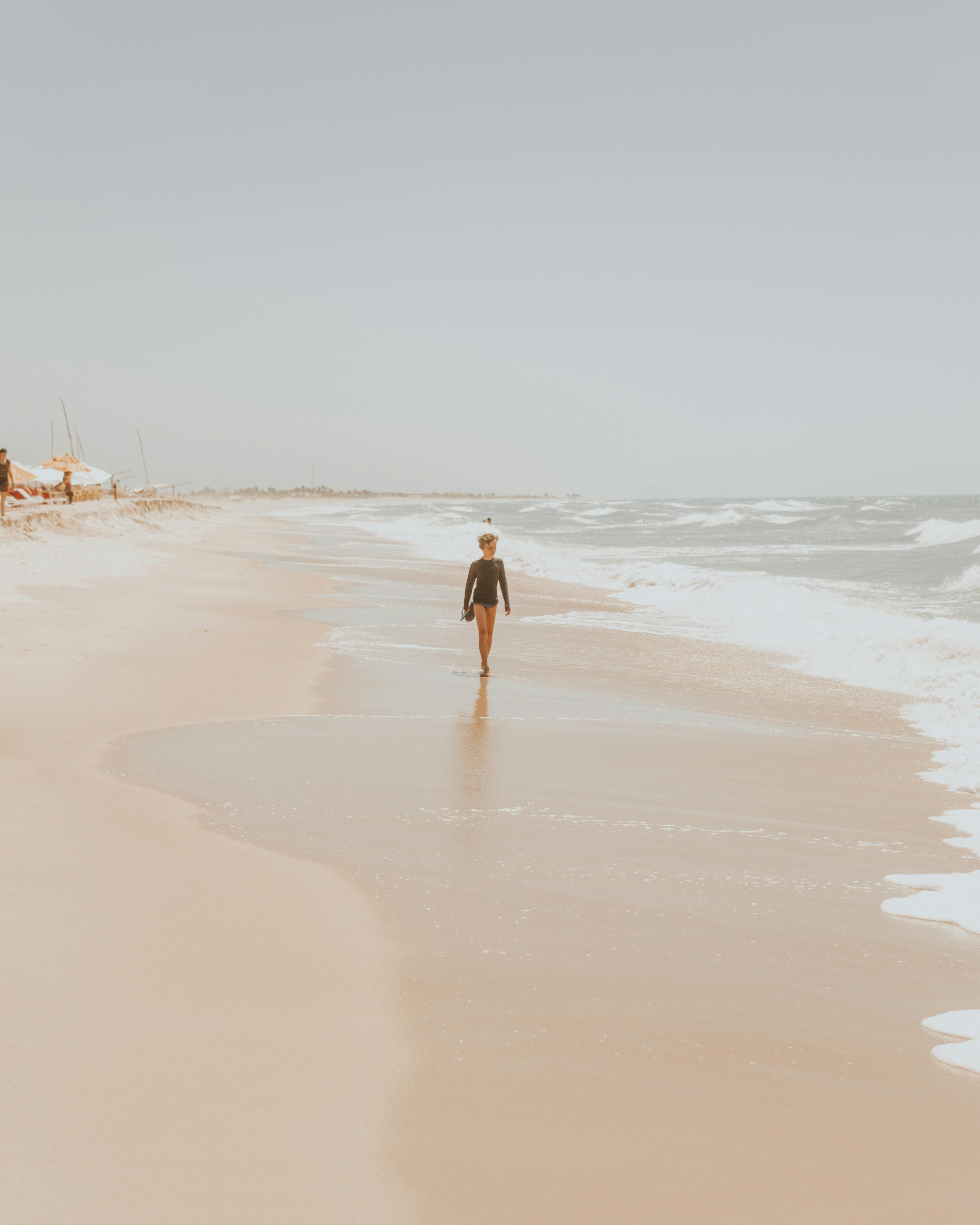 Full length faceless barefoot kid in black swimwear strolling along sandy beach washed by waving stormy sea