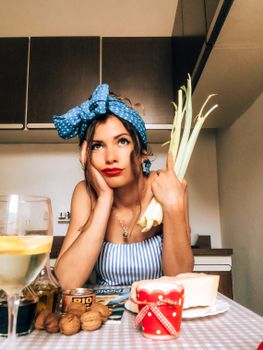 Annoyed young female wearing stylish dress and hairband sitting at table in kitchen and leaning head on hand while holding celery and looking up