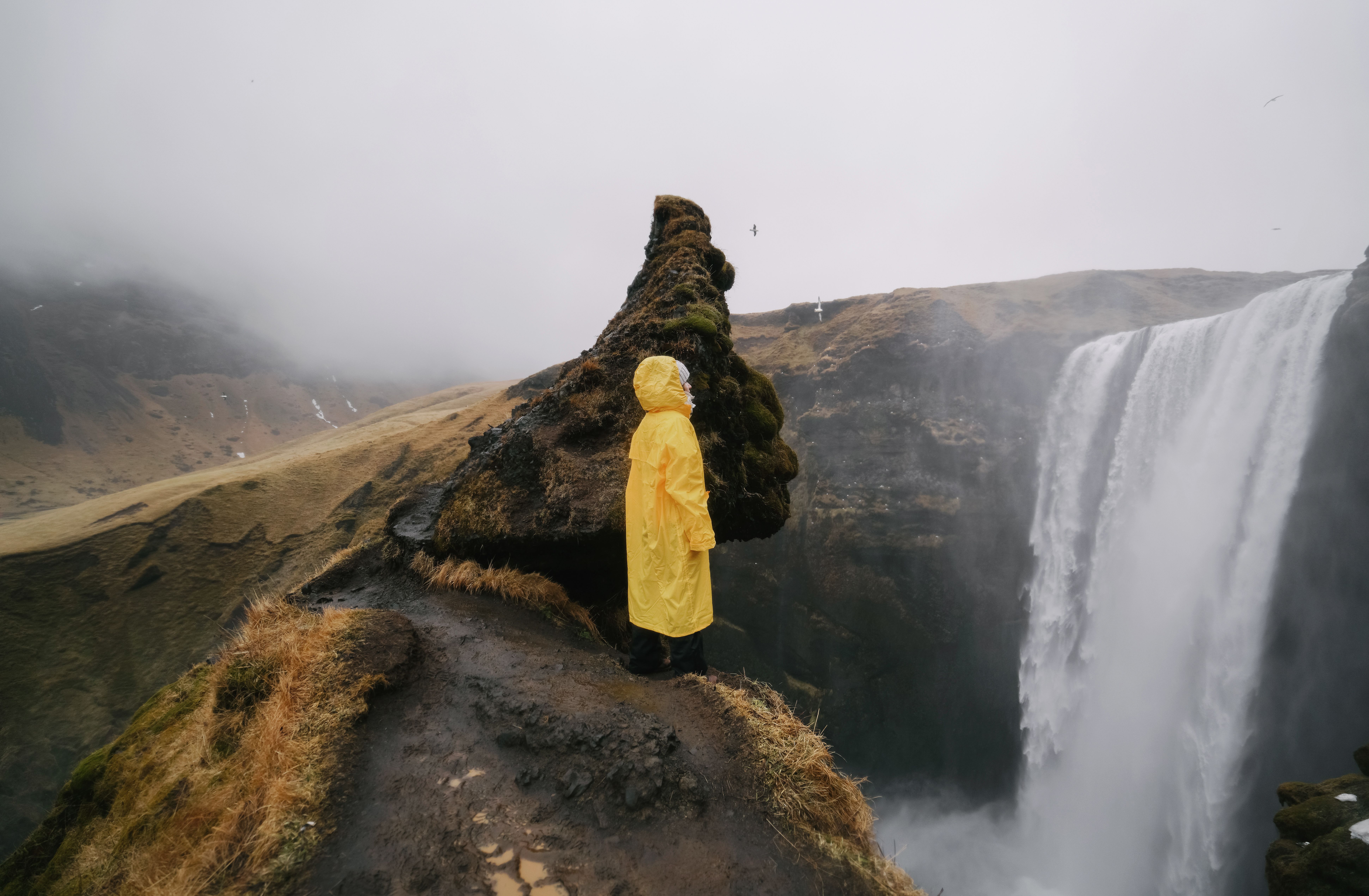 雾天里，一位身着黄色雨衣的匿名旅行者侧身立于岩石悬崖上，欣赏着山峰瀑布的如画景色