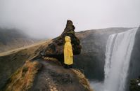 雾天里，一位身着黄色雨衣的匿名旅行者侧身立于岩石悬崖上，欣赏着山峰瀑布的如画景色