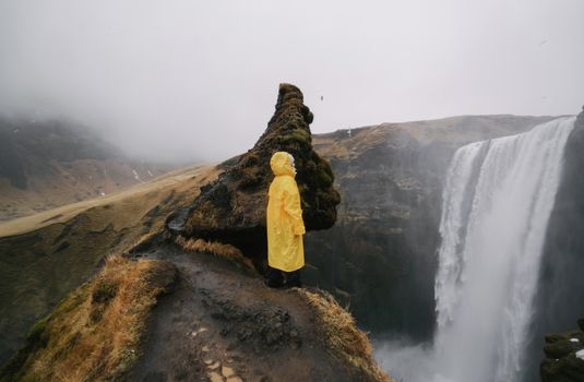 雾天里，一位身着黄色雨衣的匿名旅行者侧身立于岩石悬崖上，欣赏着山峰瀑布的如画景色