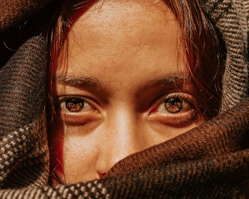 Crop unemotional female with brown eyes covering pretty face with warm brown scarf and looking at camera