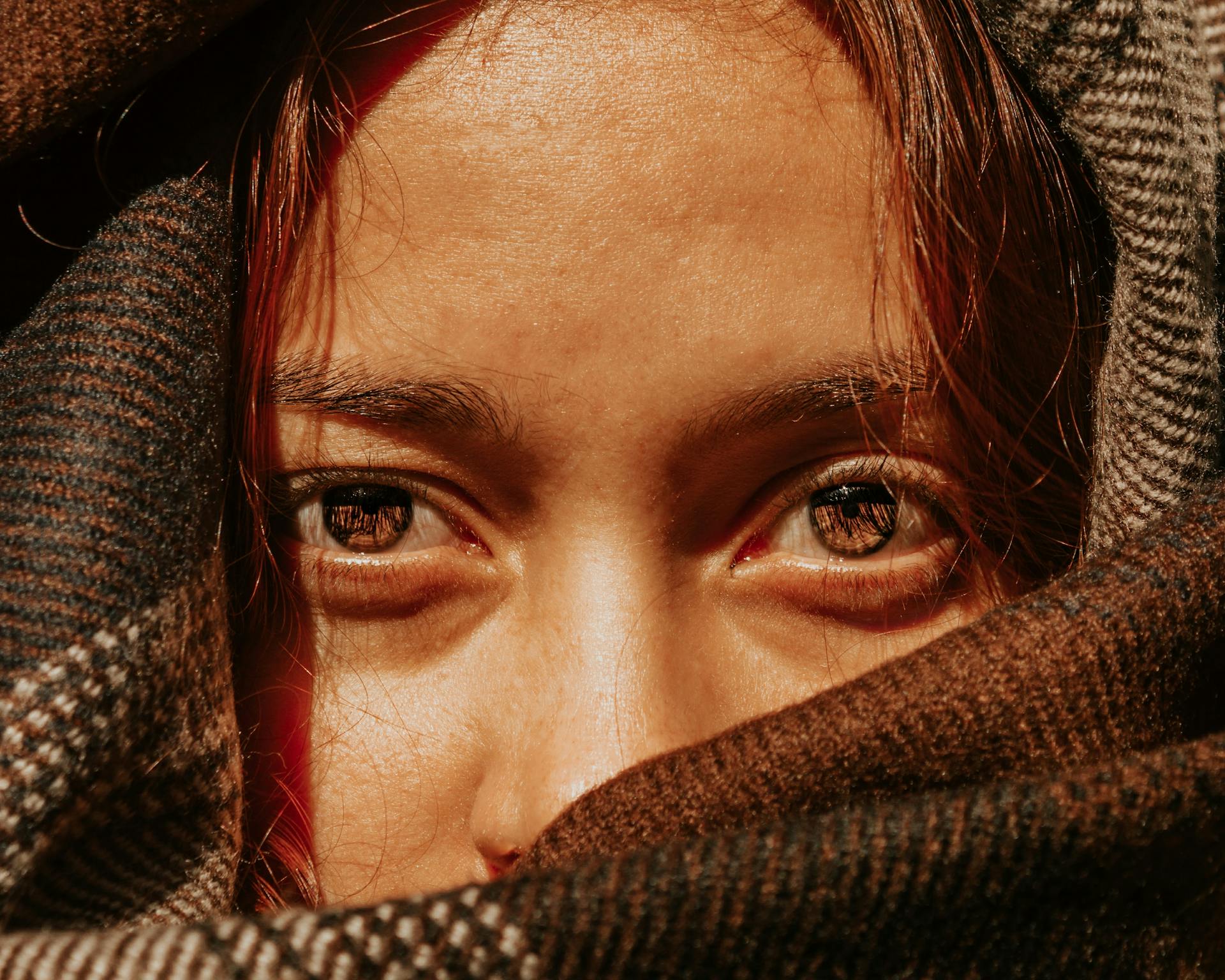 Crop unemotional female with brown eyes covering pretty face with warm brown scarf and looking at camera