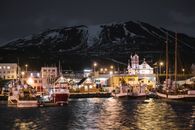 Seafront of brightly lit Husavik harbor late at night in Iceland with sailboats at quayside and dark snow covered mountain in background
