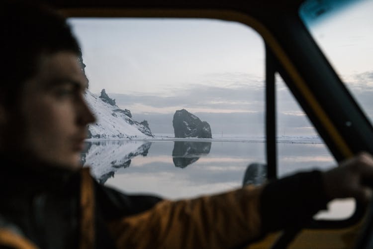 Blurry Man Driving Car Along Calm Lake And Snowy Rocks