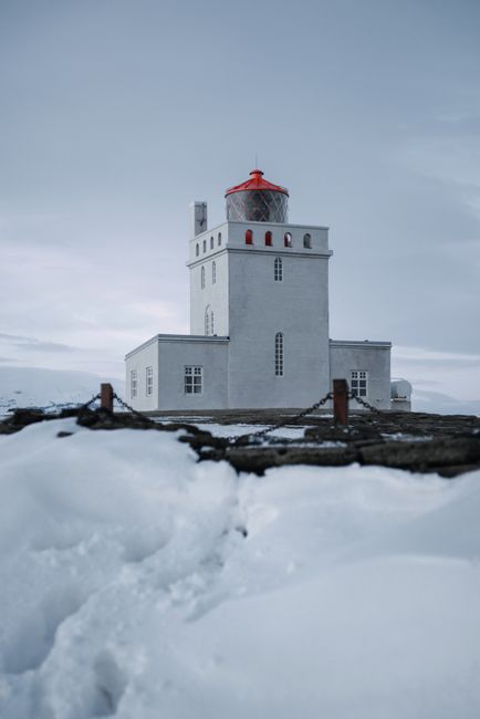 Dyrholaey Lighthouse with square concrete tower and red roof on snow covered ground under overcast sky in central south part of Iceland
